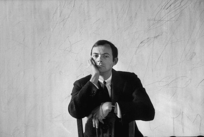 Black-and-white photograph of artist Cy Twombly from 1958, wearing a suit and tie and sitting backwards on a chair. His righthand rests on his face.