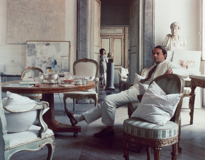 Artist Cy Twombly wearing a white suit and sitting comfortably in a chair in his art-filled apartment in a palazzo in Rome. His wife Tatiana can be seen in the doorway in the background.