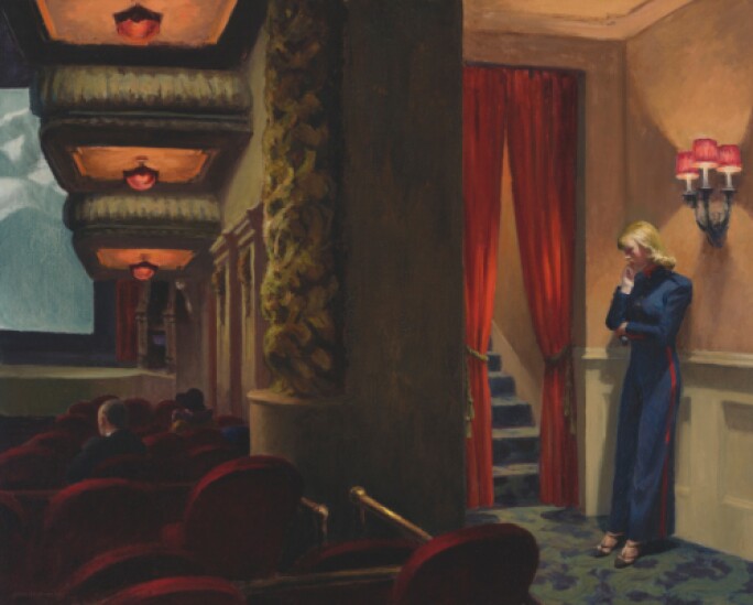 Blonde woman in a blue outfit standing alongside the dimly lit wall of a movie theater.